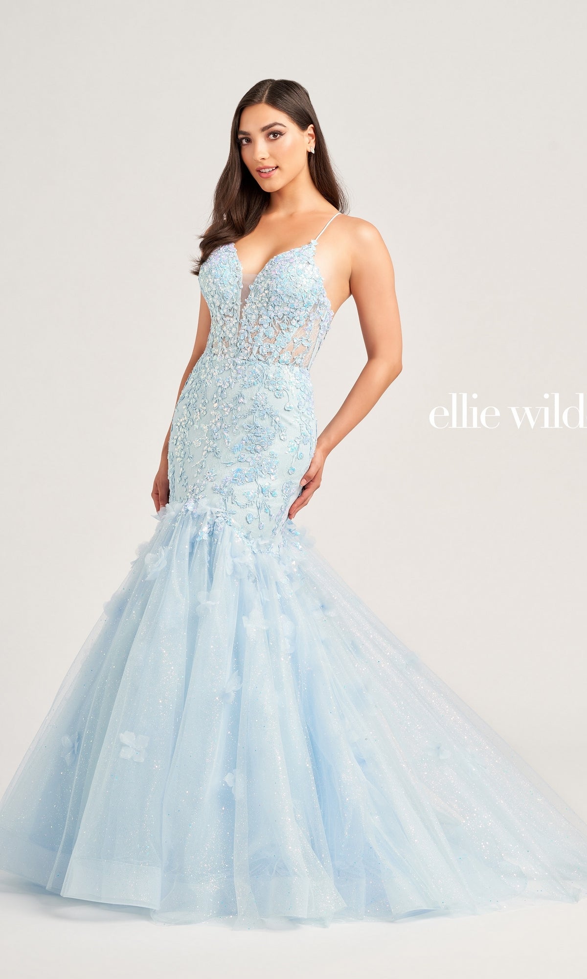A woman poses against a plain background in the Ellie Wilde Designer Mermaid Prom Dress EW35080, featuring a light blue shade, sheer bodice with floral lace appliqué, and a sparkling tulle mermaid skirt.